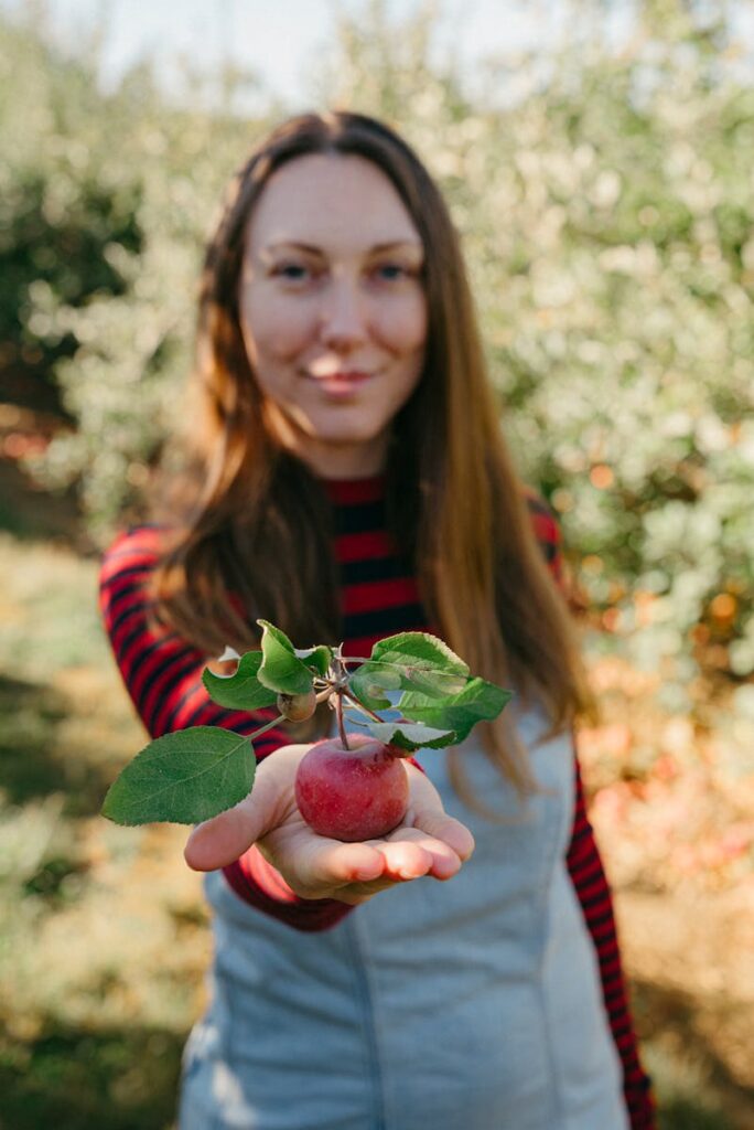 Young woman offering freshly picked red apple amidst autumn orchard.